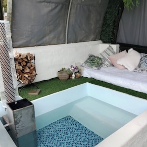 Una pequeña piscina interior con piso de mosaico azul, rodeada por un banco blanco con cojines, una pila de leña y un arreglo de plantas pequeño.