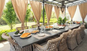 Ein Essbereich im Freien mit einem langen Tisch, gedeckt für eine Mahlzeit, umgeben von Korbstühlen und mit einer Pergola mit beigen Vorhängen.
