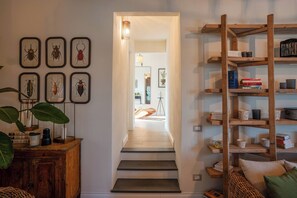 Ein Flur mit einer Holztreppe, die zu einem Zimmer mit einem Bett und einem Schreibtisch führt. An der Wand befinden sich gerahmte Insektenbilder und ein Holzregal mit Büchern und Dekorationsobjekten.