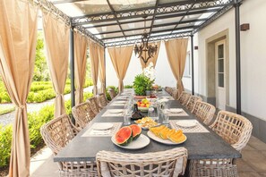An outdoor dining area with a long table set for a meal, surrounded by wicker chairs, and a pergola with curtains. - Villa Asti a luxury villa for enjoyment in Tuscany (Capannori)