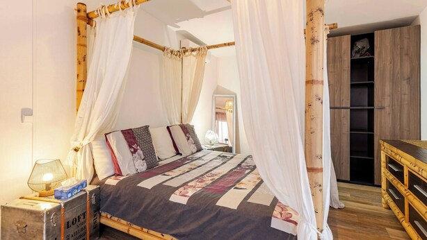 A four-poster bed with a floral bedspread, a bedside lamp, and a wooden nightstand with a suitcase. - Vrbo Property (Saint-Louis)