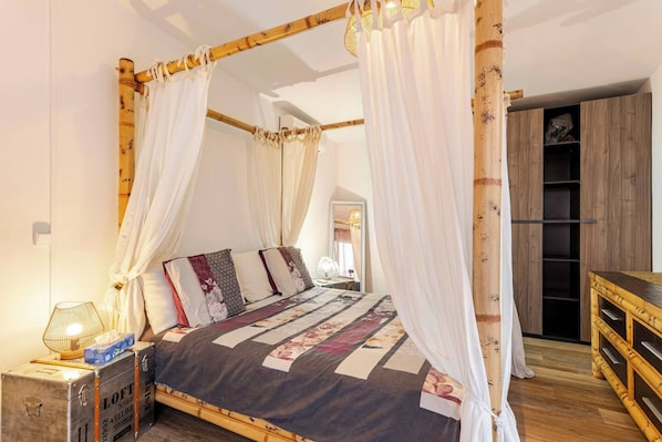 Un lit à baldaquin avec une couvre-lit à motifs floraux, une lampe de chevet et une table de chevet en bois sur laquelle repose une valise.