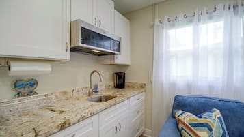 Una cocina con armarios blancos, un lavamanos de acero inoxidable, una campana extractora integrada y un ventanal con cortinas traslúcidas.