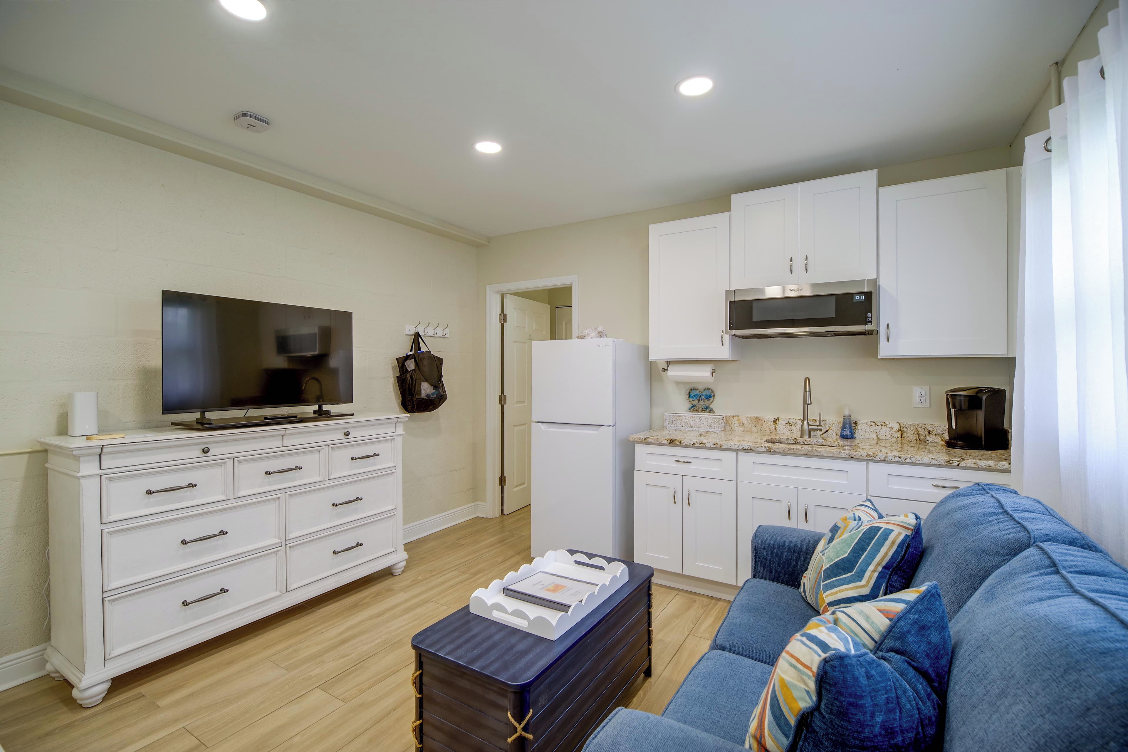 Una sala de estar moderna con un televisor de pantalla plana, un sofá, una mesa de centro y una zona de cocina con armarios blancos y un microondas.