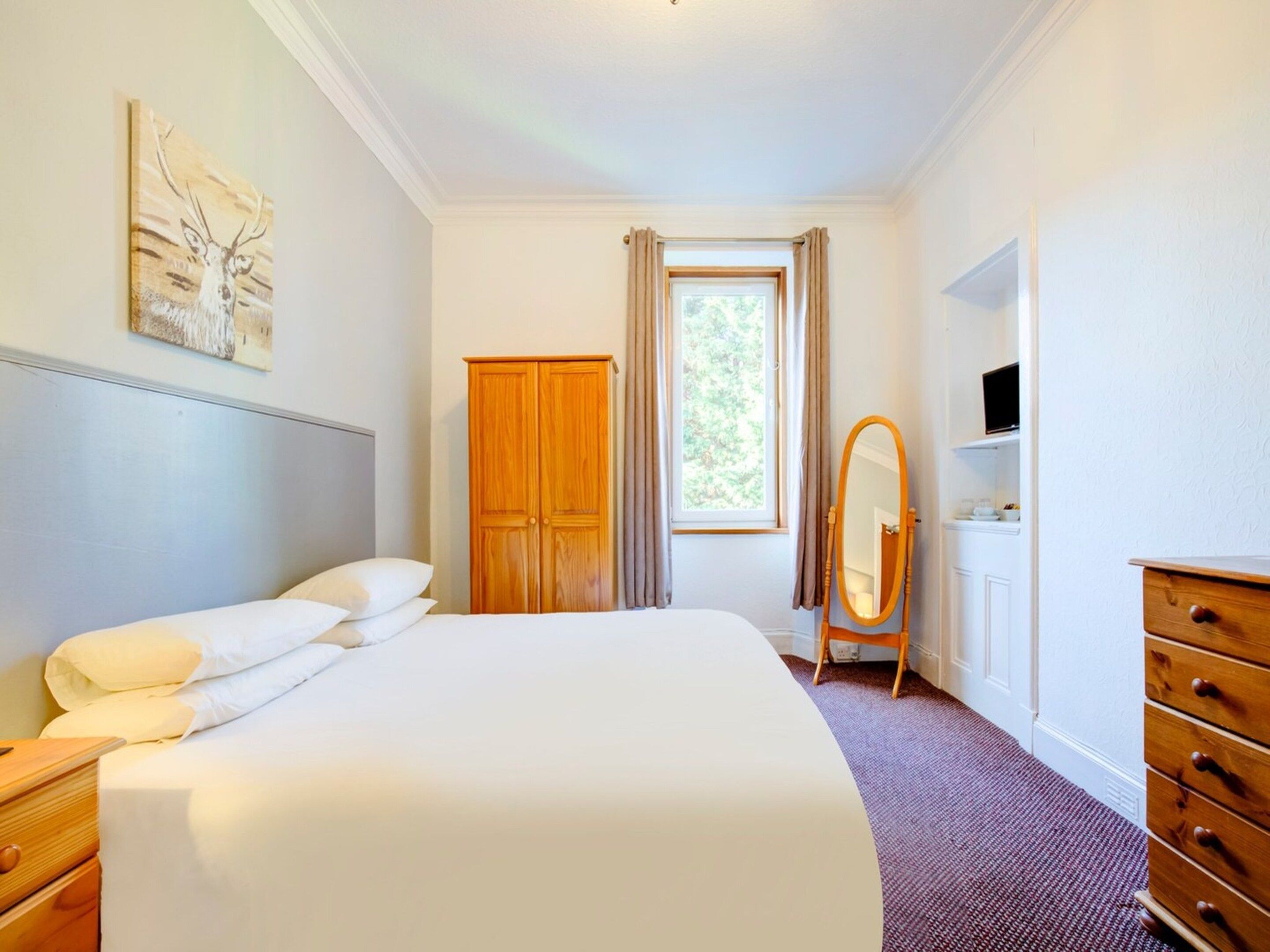 A hotel room with a bed, a wooden wardrobe, a dressing table with a mirror, a television on the wall, and a window with curtains.