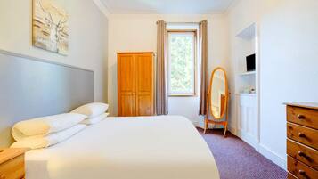 A hotel room with a bed, a wooden wardrobe, a dressing table with a mirror, a television on the wall, and a window with curtains.