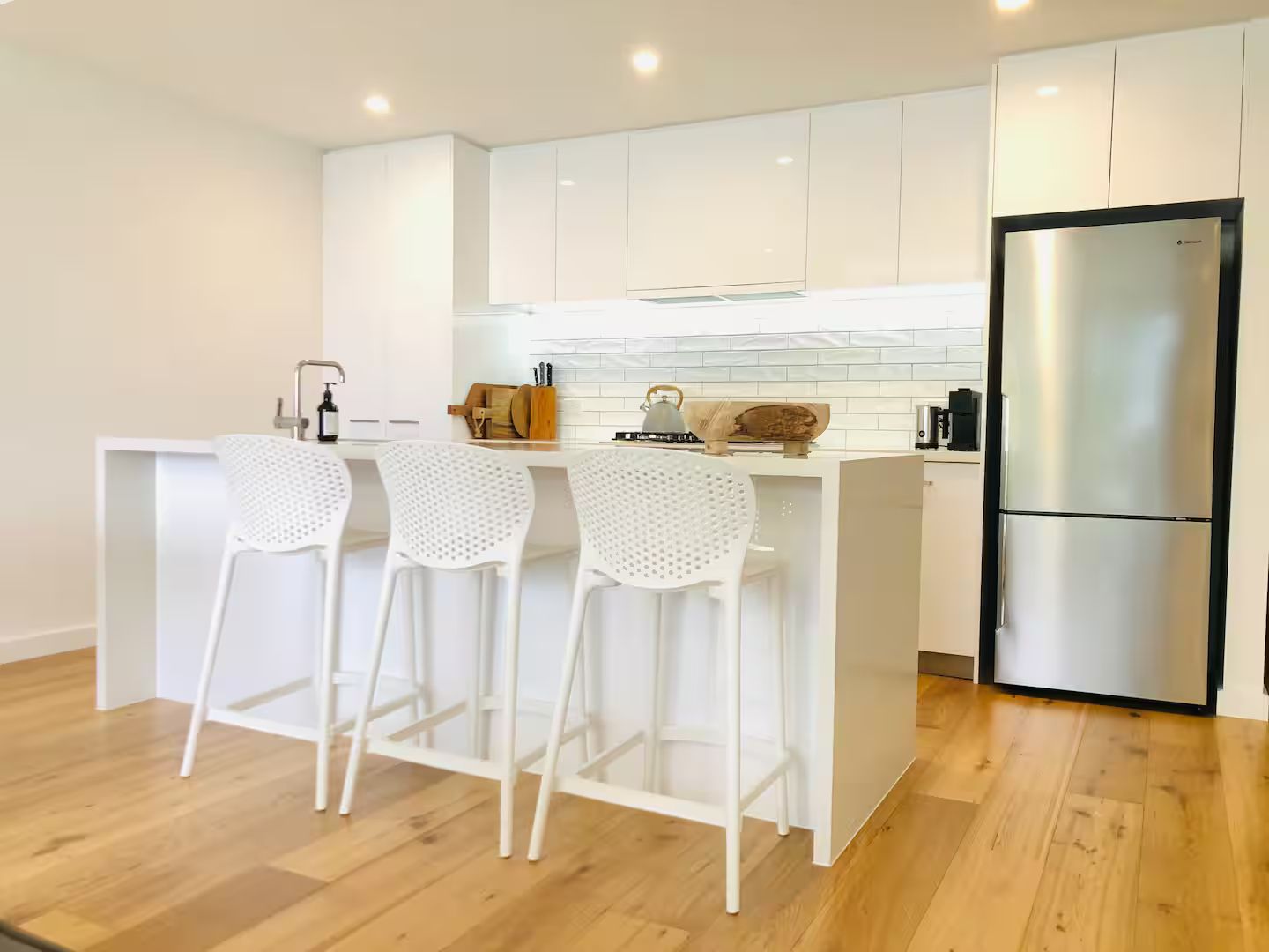 A modern kitchen with white cabinetry, a stainless steel refrigerator, and three white bar stools.