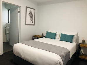 Un dormitorio con una cama, dos mesitas de noche, una mesita auxiliar, una obra de arte enmarcada y una puerta que da a un baño.