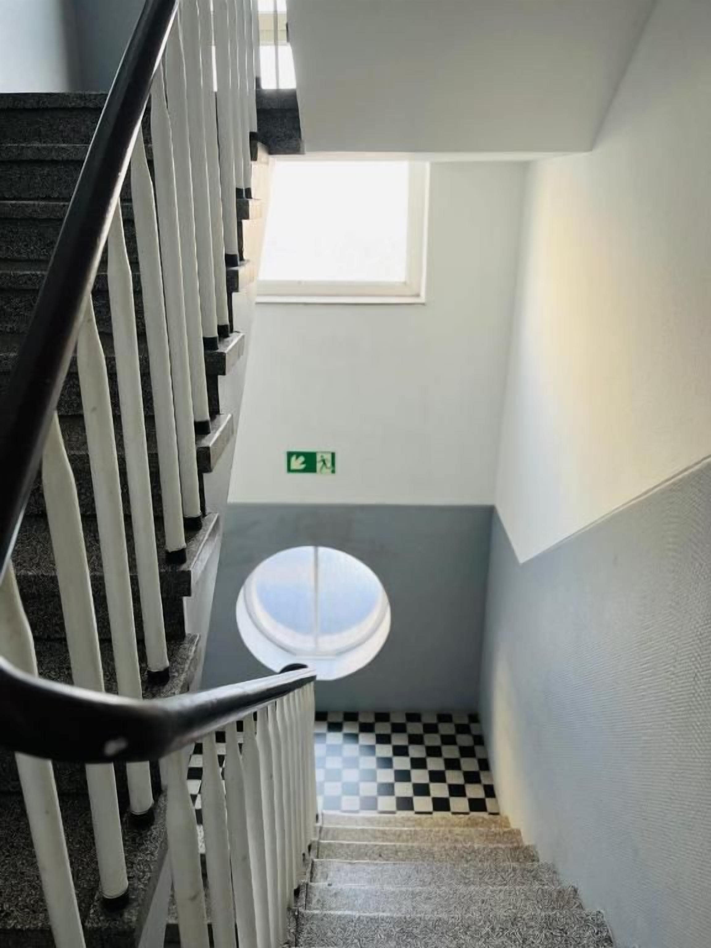 Eine Treppe mit einem Schachbrettmuster im Boden, einer weißen Brüstung und einem kleinen Fenster am oberen Ende.