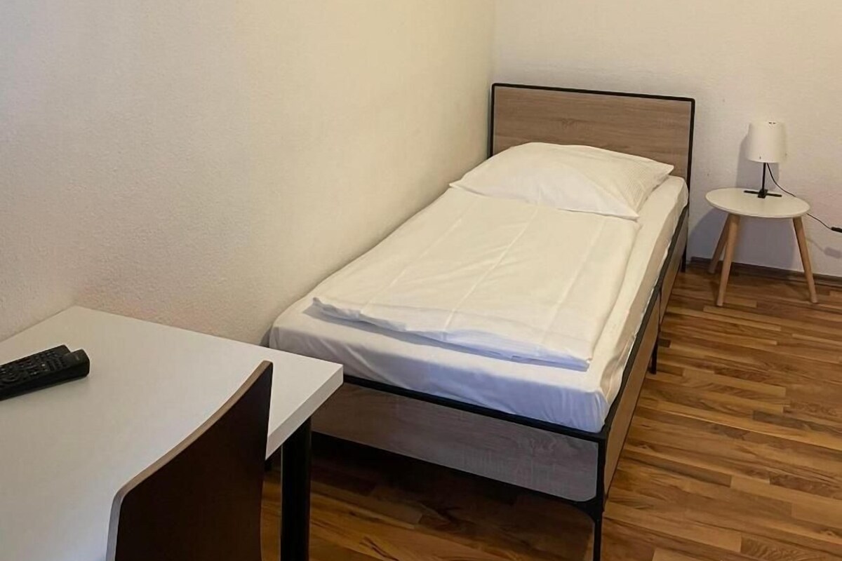 A single bed with a wooden headboard, a small bedside table with a lamp, and a desk with a remote control.