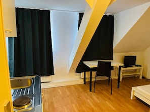 Una habitación pequeña y triangular con kitchenette, una cama individual, un escritorio con silla y televisión.
