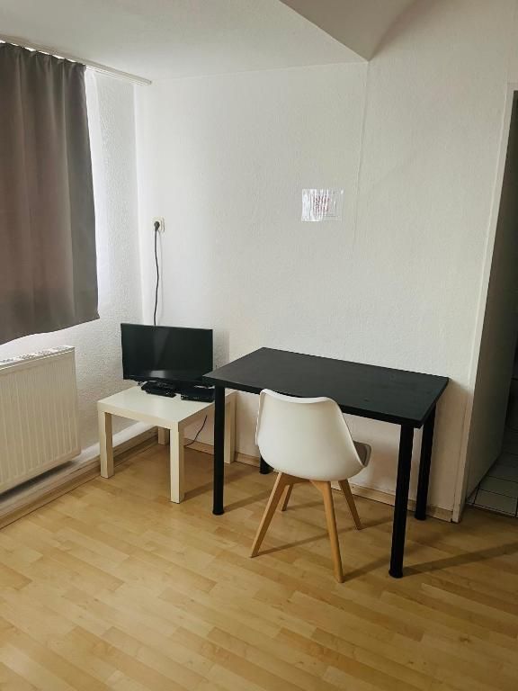 Ein Zimmer mit einem schwarzen Tisch, einem weißen Stuhl, einem Fernseher auf einem Schrank und einem Fenster mit Vorhängen.