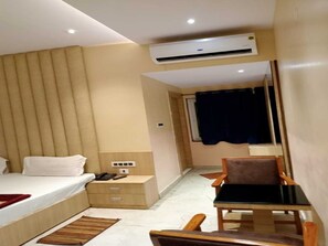 A hotel room with a bed, bedside table, chair, and an air conditioning unit.