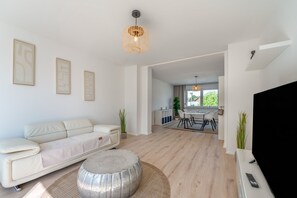 Ein modernes Wohnzimmer mit einer Couch, einem Couchtisch und einem Fernseher. Im Hintergrund ist ein Essbereich mit Tisch und Stühlen zu sehen.