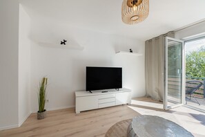 A modern living room with a flat-screen TV on a white cabinet, a potted plant, and a large window with a view of greenery. - Apartment "Onjito" with Shared Garden, Balcony, and Wi-Fi (Saarbrücken)