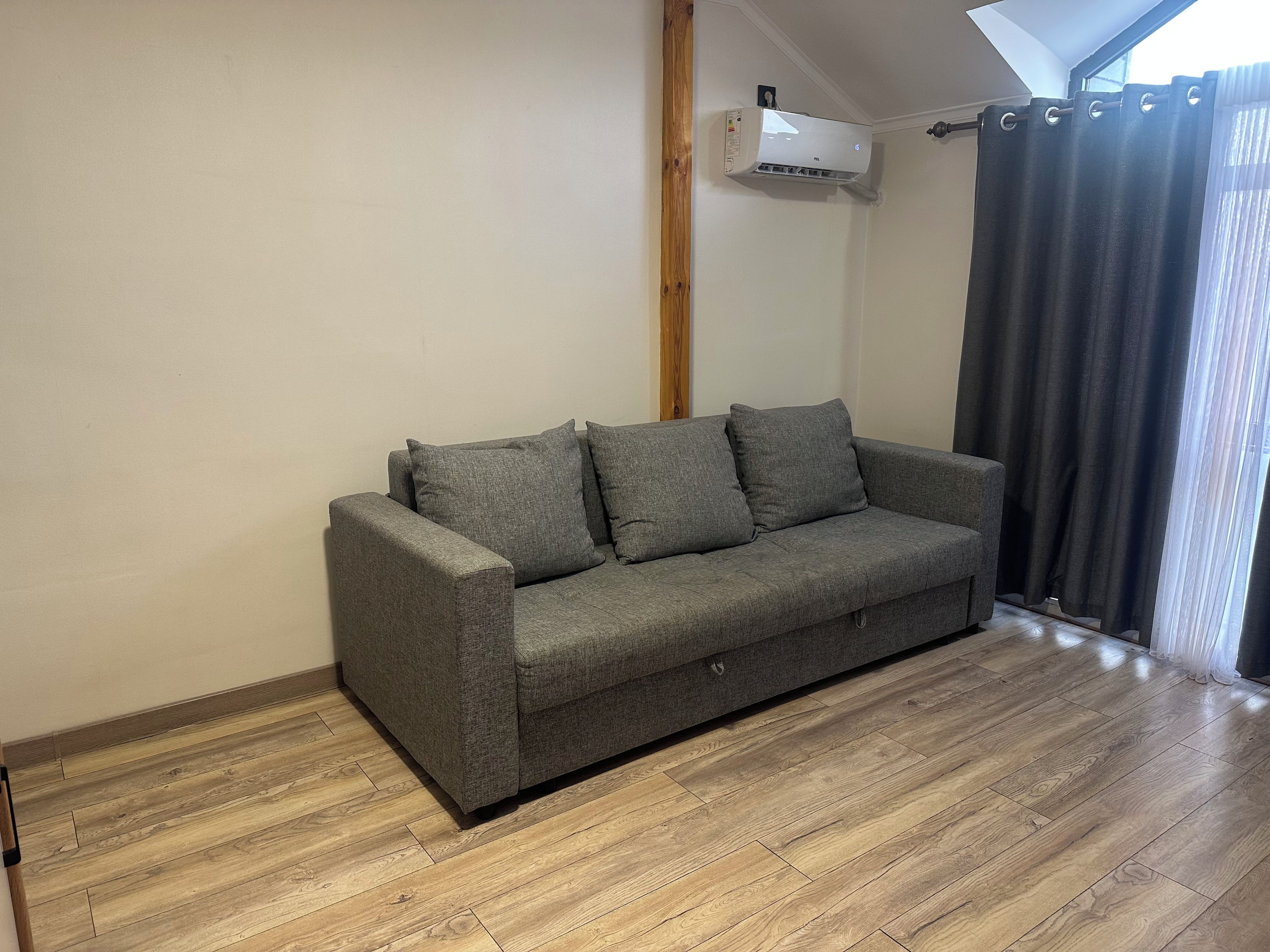 A grey sofa in a room with wooden flooring, a window with curtains, and an air conditioning unit mounted on the wall.