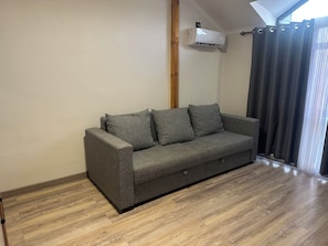 A grey sofa in a room with wooden flooring, a window with curtains, and an air conditioning unit mounted on the wall.