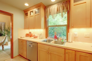 A kitchen with wooden cabinets, a dishwasher, a window with curtains, and various kitchen items on the counter. - Hike, Ski & Soak! Mt. Hood Getaway w/ Hot Tub (Rhododendron)