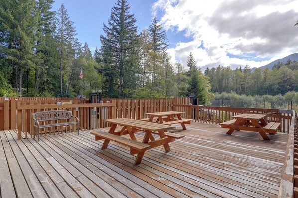 Eine Holzterrasse mit Picknicktischen und einer Bank, umgeben von einem Holzzaun und hohen Bäumen.