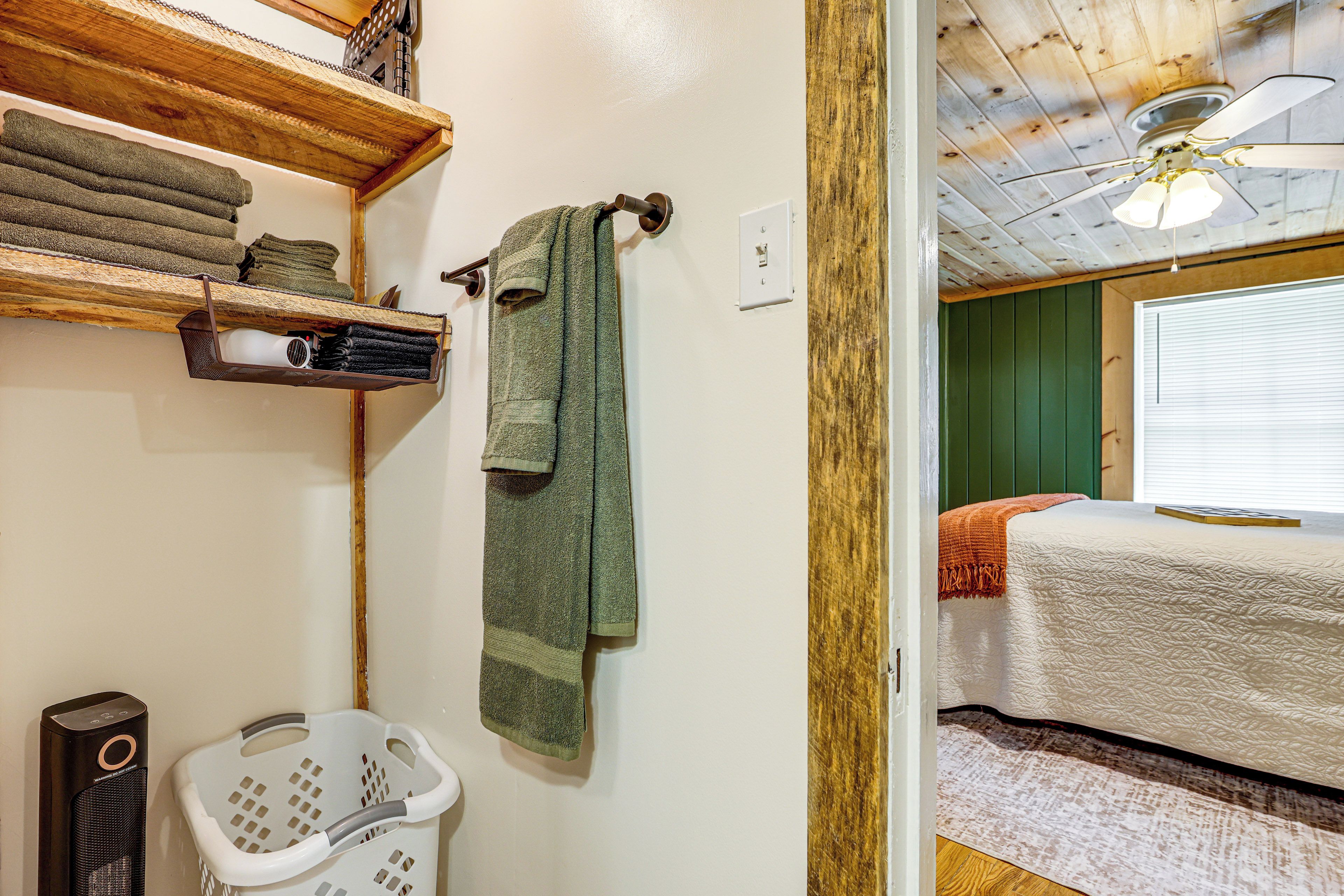 A bathroom with a wooden shelf holding towels, a hanging towel, a white laundry basket, and a ceiling fan.