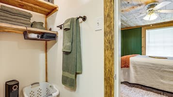 A bathroom with a wooden shelf holding towels, a hanging towel, a white laundry basket, and a ceiling fan.