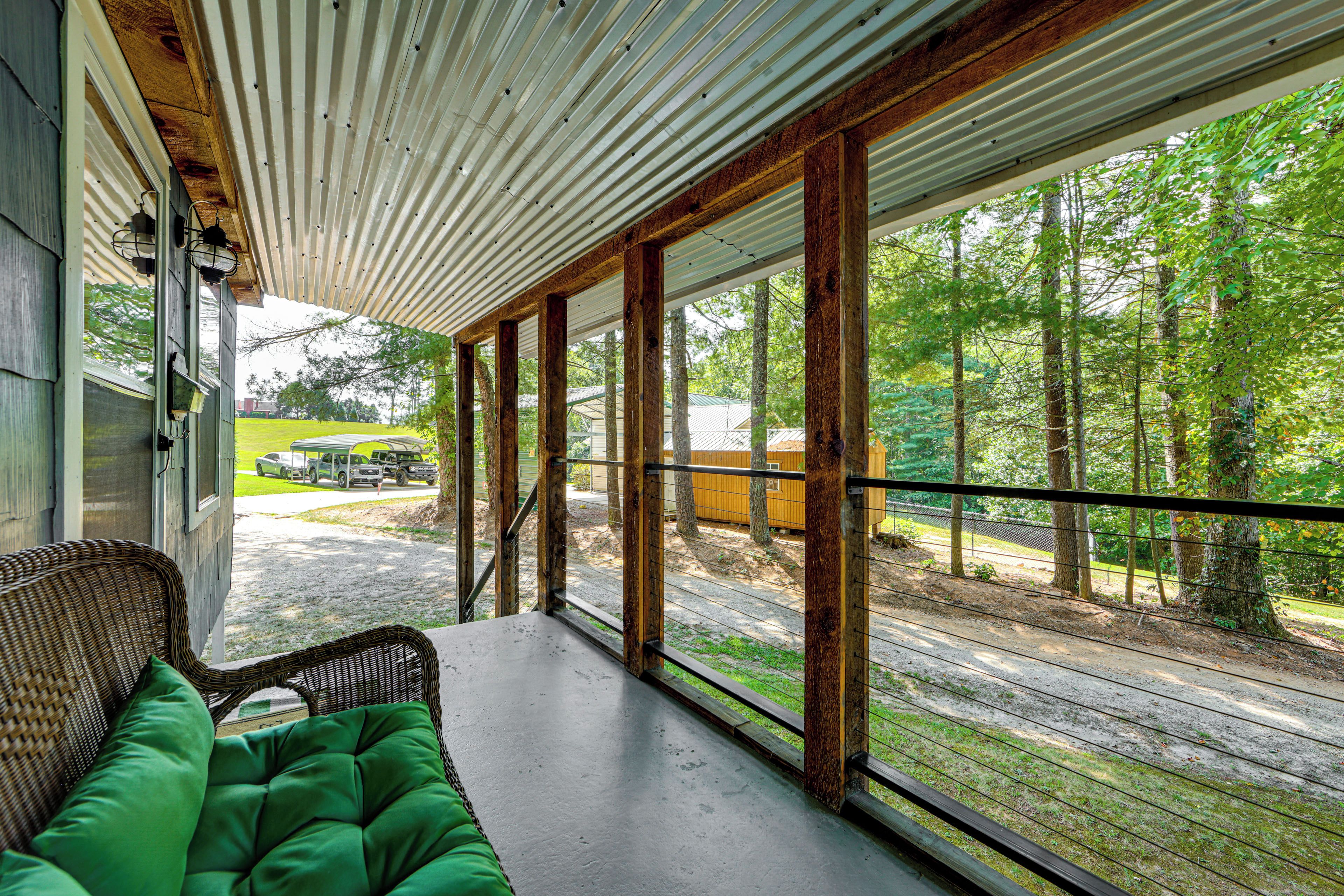 A covered porch with a wicker chair and a green cushion, overlooking a forested area with a gravel path and parked vehicles.