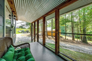 A covered porch with a wicker chair and a green cushion, overlooking a forested area with a gravel path and parked vehicles. - Cozy Candler Cottage, Near Hiking and Wildlife (Candler)