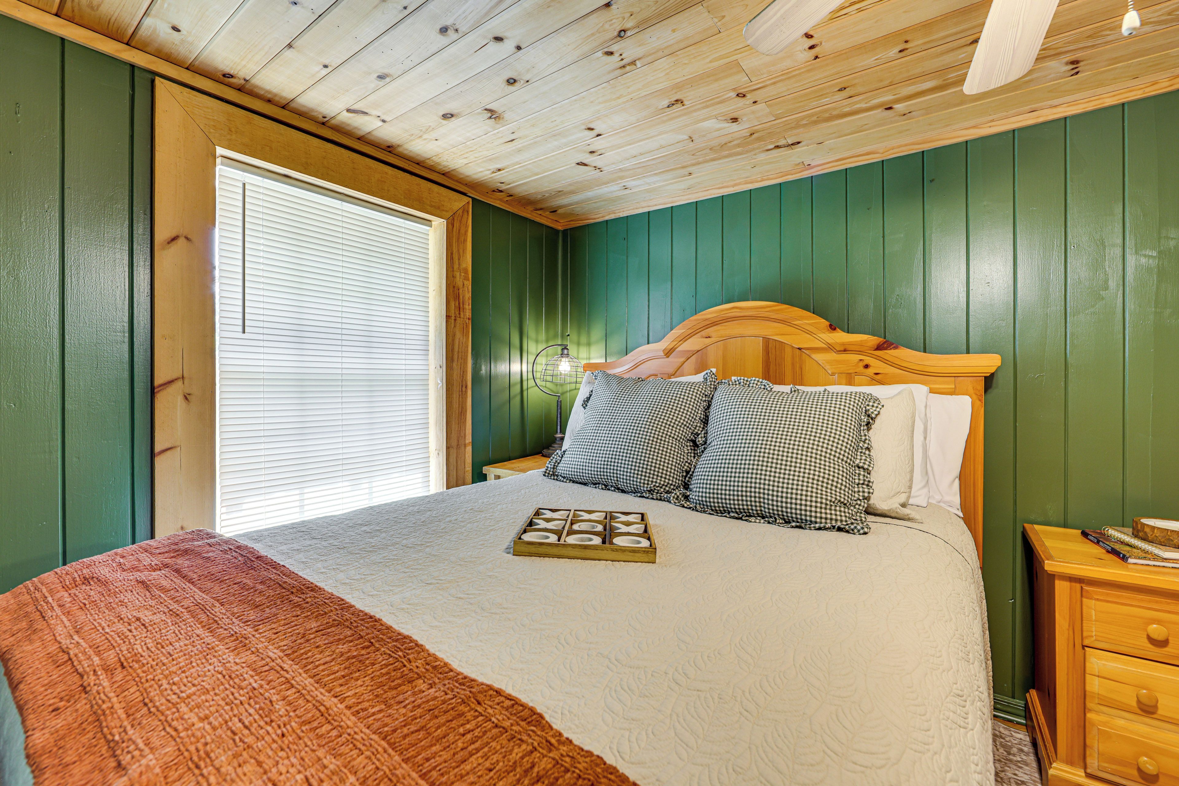 A bedroom with a wooden headboard, a bed with a white comforter, checkered pillows, a wooden nightstand, and a window with blinds.