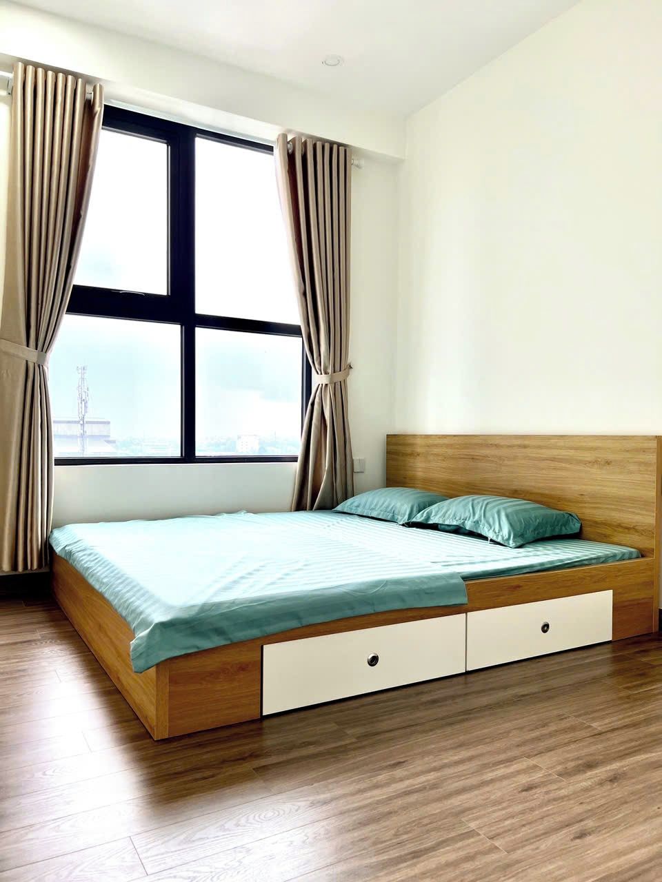 Quarto com cama de madeira, janela com cortinas e vista para a paisagem urbana.