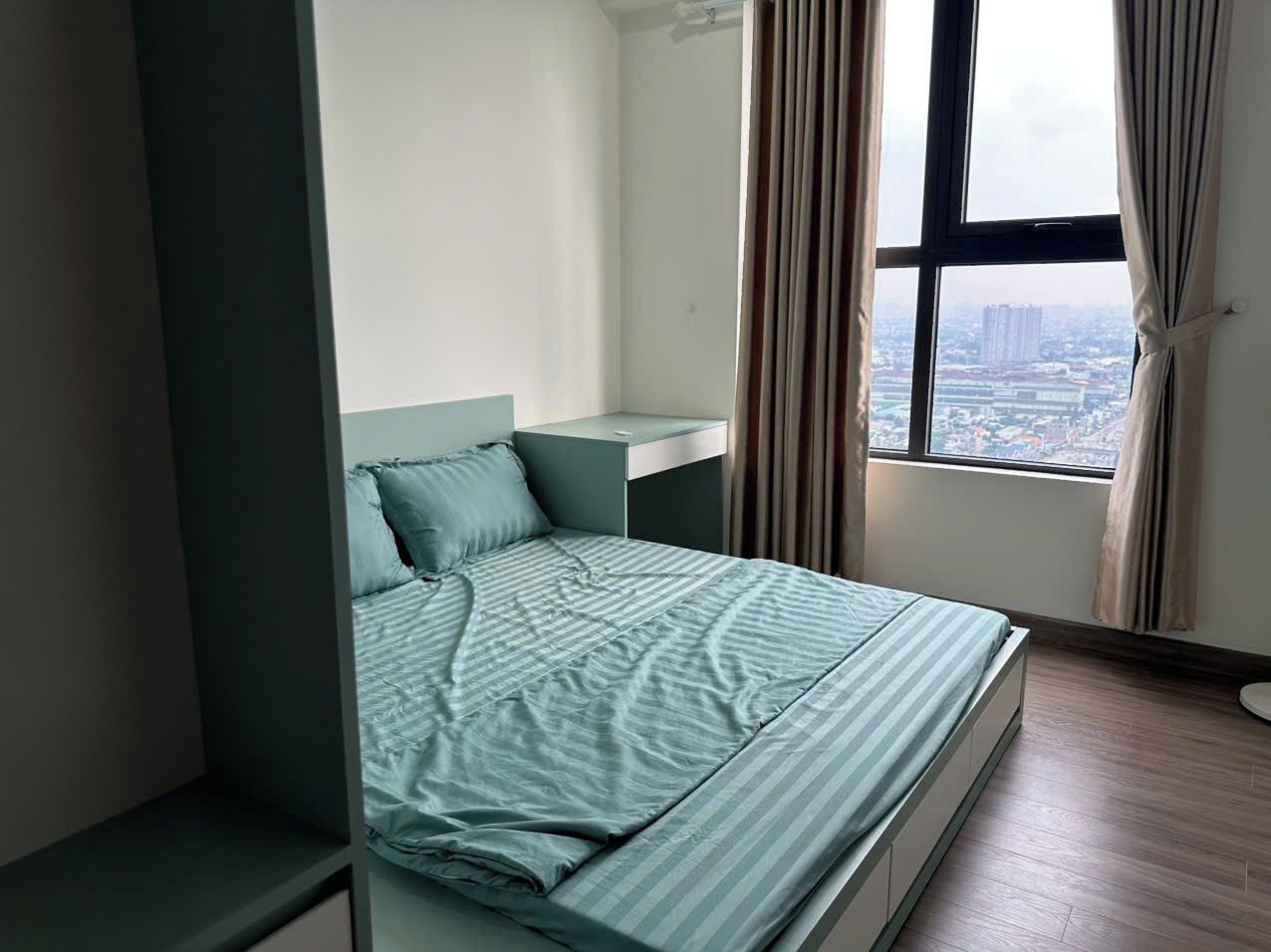 Uma cama de solteiro com cabeceira, mesa de cabeceira e vista para a paisagem urbana através de uma janela com cortinas.