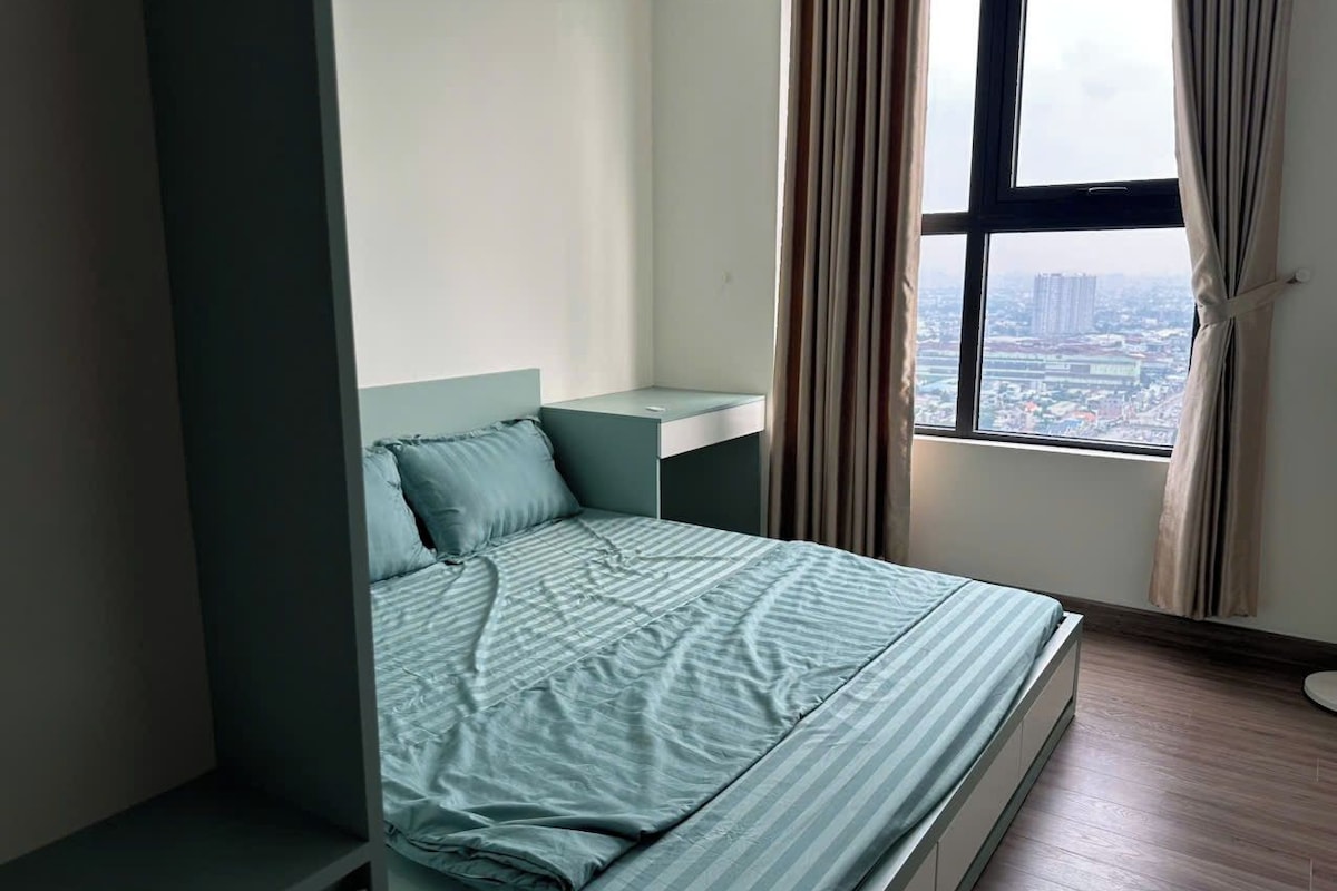 Uma cama de solteiro com cabeceira, mesa de cabeceira e vista para a paisagem urbana através de uma janela com cortinas.