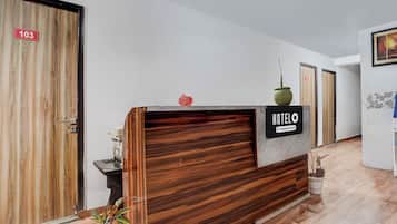 Un mostrador de recepción de hotel con detalles de madera, un letrero que indica que es un hotel con servicios de empresa, y un área de recepción con una planta en maceta.