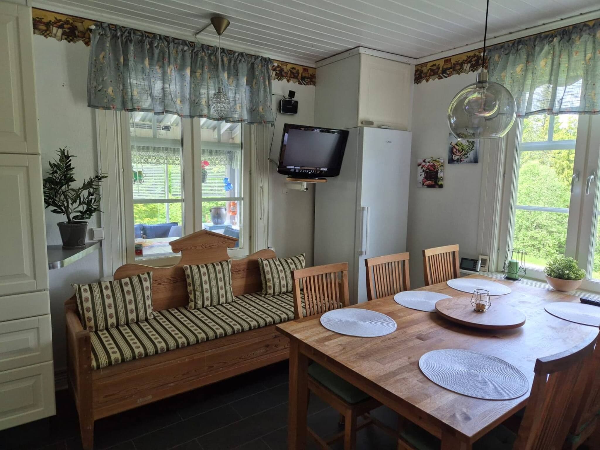 A cozy room with a wooden sofa, a dining table with placemats, a television mounted on the wall, and a window with curtains.