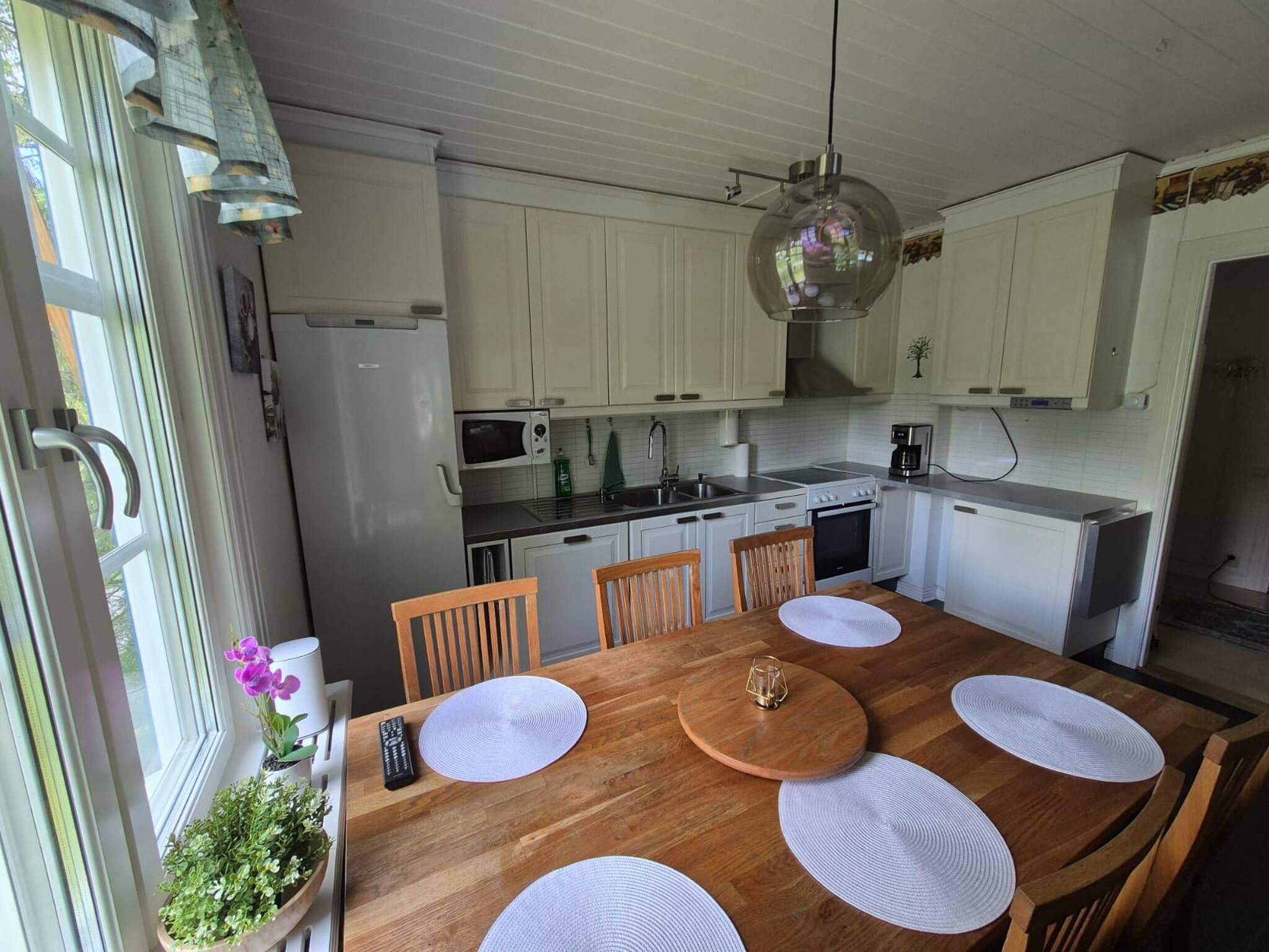 A kitchen with white cabinets, a wooden dining table with placemats, and a hanging light fixture.
