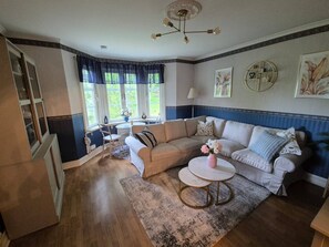 A living room with a beige sofa, a coffee table, and a patterned rug. There is a window with curtains, a lamp, and wall art. - Holiday House With Lots of Charm (Lunde)