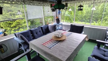 A covered outdoor dining area with a table and chairs, surrounded by windows with blinds, and a view of greenery outside.