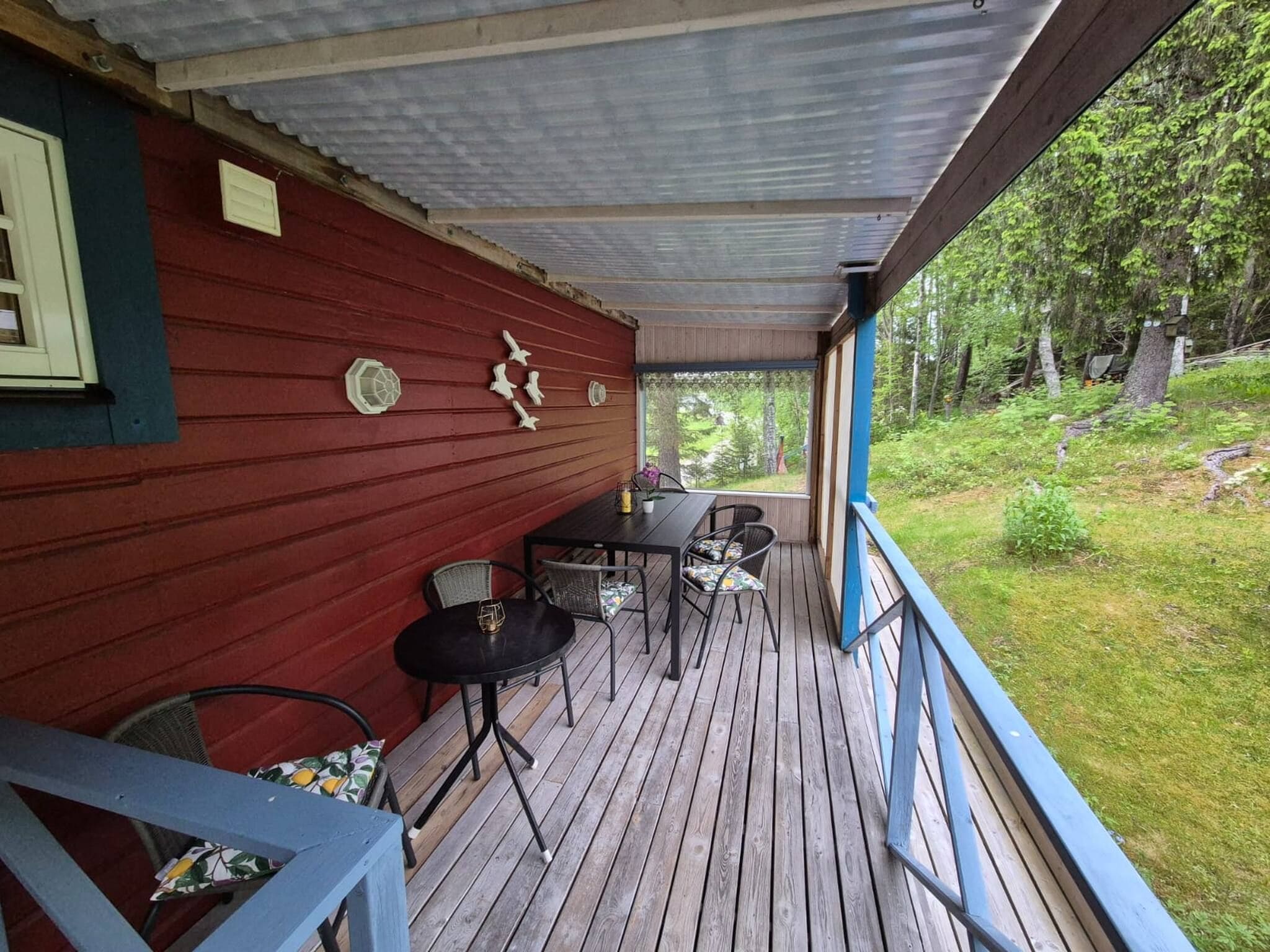 A covered wooden deck with a red wall, a table with chairs, and a view of the surrounding greenery.
