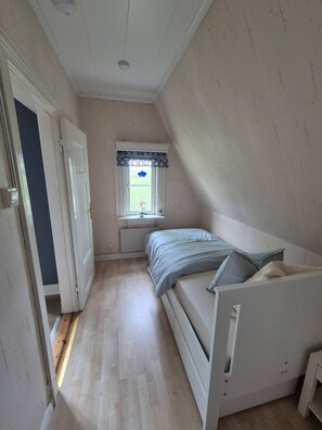 Habitación pequeña con cama individual, techo inclinado, ventana con un adornado cortinero y puerta que da a otra habitación.