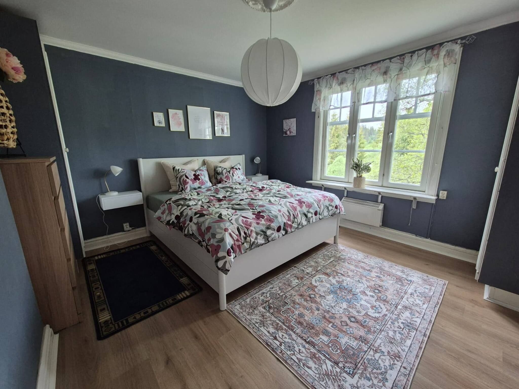 A bedroom with a bed, bedside tables, a rug, a window with curtains, and wall art.