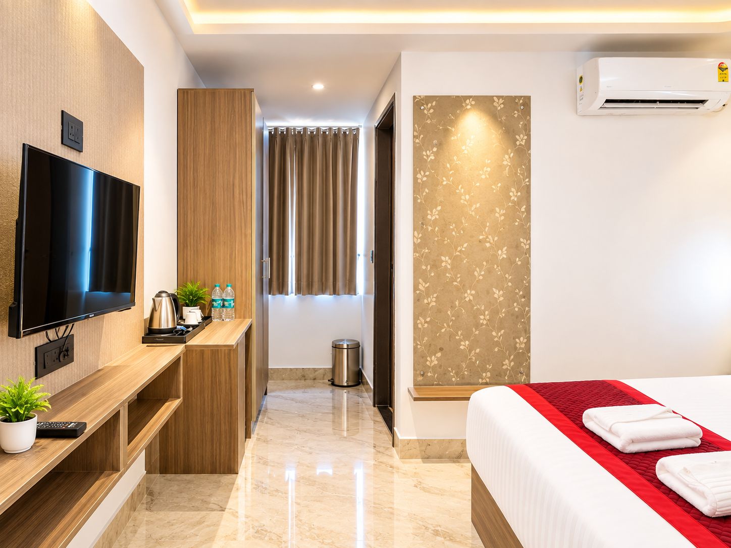 A hotel room with a flat-screen TV, a wooden desk, a bed with a red accent, and an air conditioning unit.