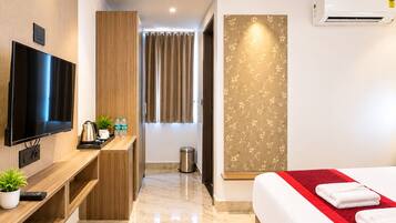 A hotel room with a flat-screen TV, a wooden desk, a bed with a red accent, and an air conditioning unit.