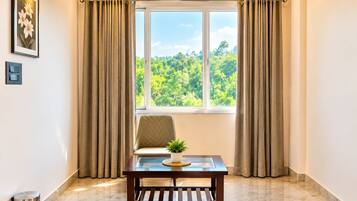 A room with a large window, a cushioned chair, a small table with a plant, and a trash bin.