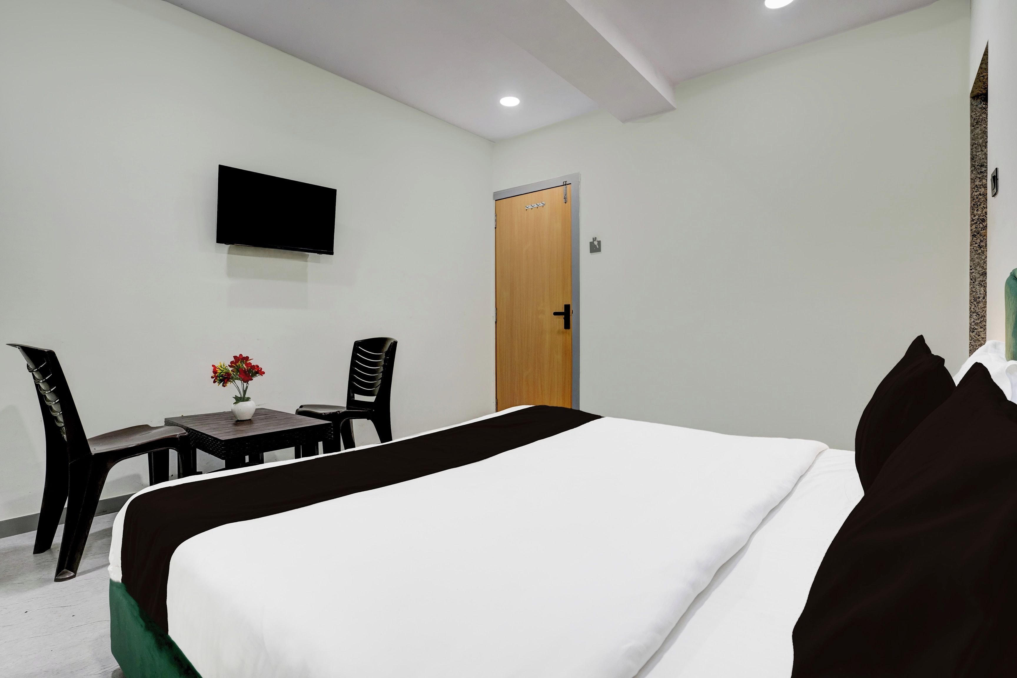 Una habitación de hotel con cama, televisión montada en la pared, puerta de madera y una mesita con un florero.