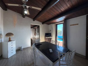 A room with a wooden ceiling, a dining table with chairs, a television, and a view of the sea through a window. - Il Cigno Reale White (Chiaramonte Gulfi)