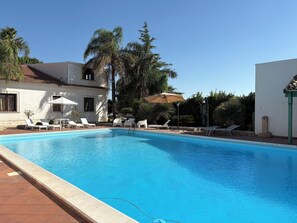A swimming pool with lounge chairs and umbrellas, surrounded by palm trees and a building with a tiled roof. - Il Cigno Reale White (Chiaramonte Gulfi)