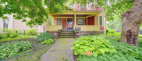 A two-story house with a porch, a front yard with various plants, and a large tree.