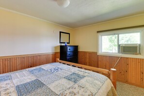 A bedroom with a wooden headboard, a quilted bedspread, a dresser, a mirror, and a window with an air conditioning unit. - Fire Pit & Views! Lake Champlain Shoreline Stay (Plattsburgh)