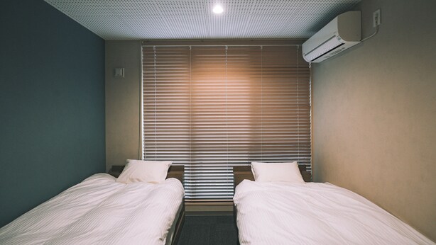 A hotel room with two single beds, a window with blinds, a ceiling with a grid pattern, and an air conditioning unit on the wall. - Ted's guesthouse (Uchiko)