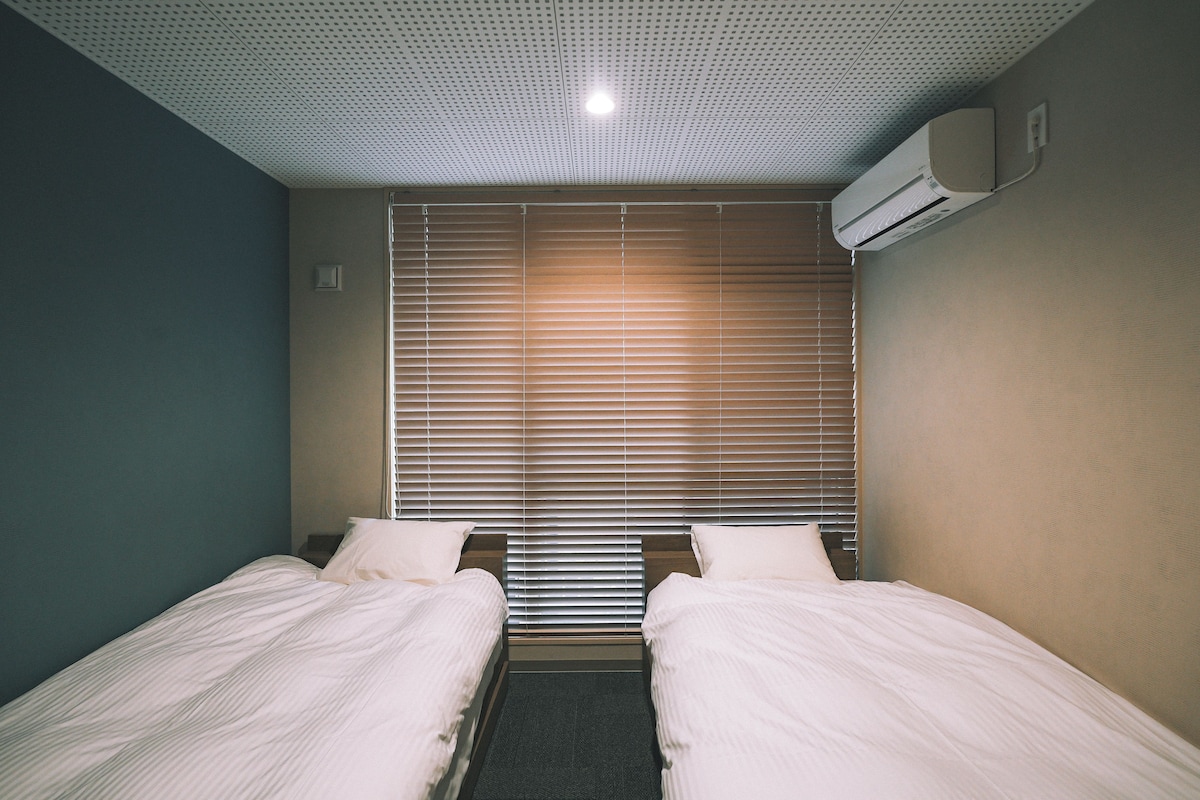 A hotel room with two single beds, a window with blinds, a ceiling with a grid pattern, and an air conditioning unit on the wall.