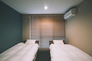 A hotel room with two single beds, a window with blinds, a ceiling with a grid pattern, and an air conditioning unit on the wall. - Ted's guesthouse (Uchiko)